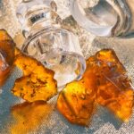 Several pieces of amber, translucent cannabis shatter beside a clear glass jar on a textured metallic surface