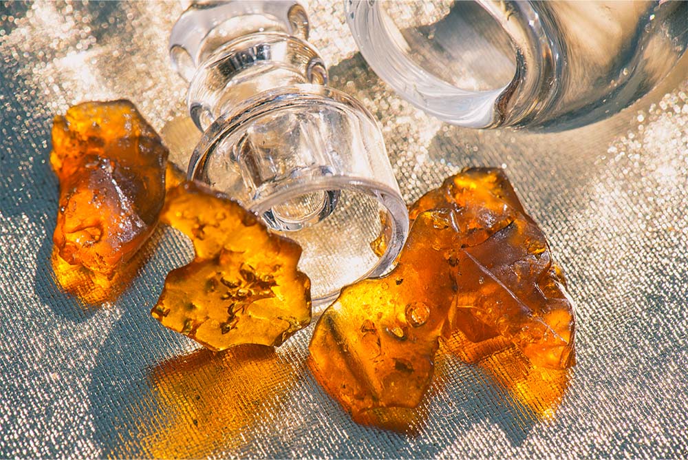Several pieces of amber, translucent cannabis shatter beside a clear glass jar on a textured metallic surface